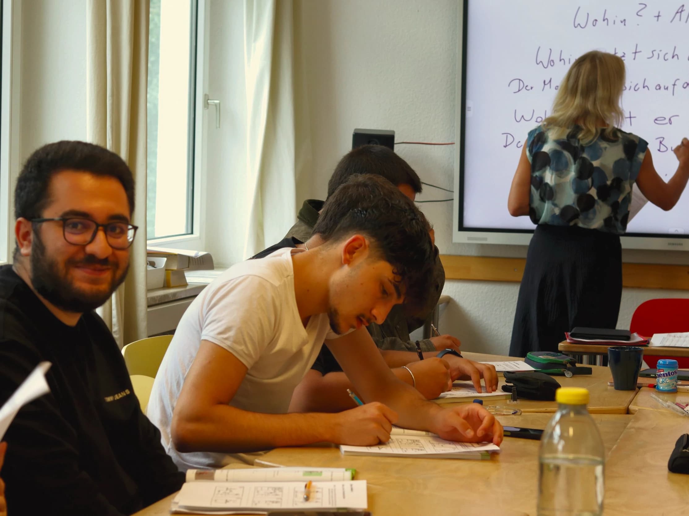 Ein Teilnehmer lächelt von seiner Übung auf, die Lehrerin schreibt Grammatik an die Tafel