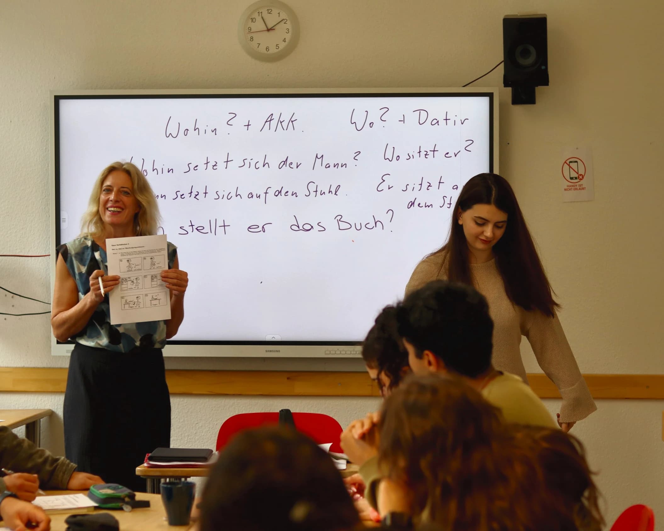 Eine Lehrerin hält lachend ein Arbeitsblatt hoch, an der Tafel stehen die Wechselpräpositionen